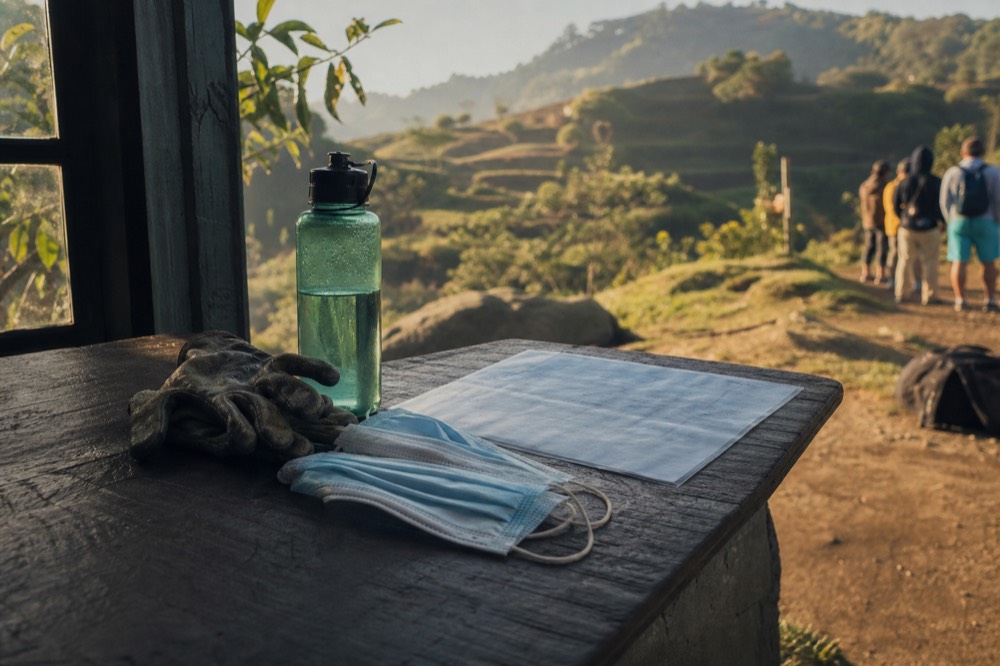 A pair of surgical face masks, a water bottle and gardening gloves laid on a park briefing table beside a laminated rules card, morning light through a window