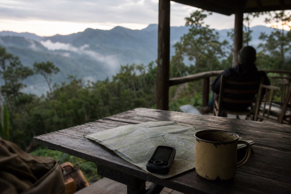 A weathered field map and a battered GPS unit resting on a wooden table beside a chipped enamel mug, montane forest visible beyond the veranda rail
