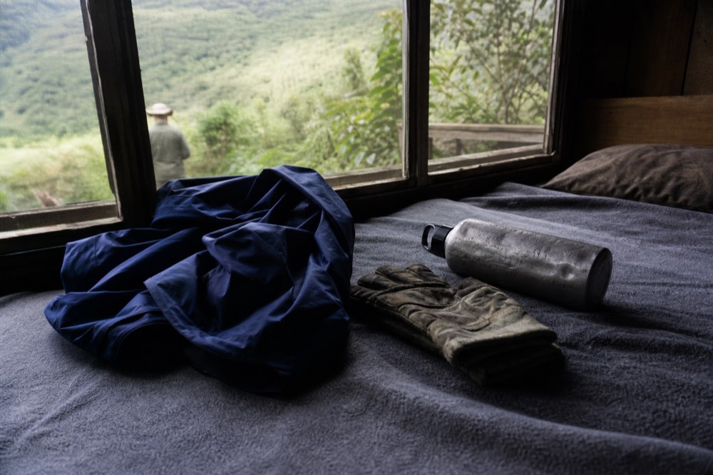A folded rain shell, worn gardening gloves and a dented water bottle laid out on a lodge bed before an early start, cool window light across the blanket