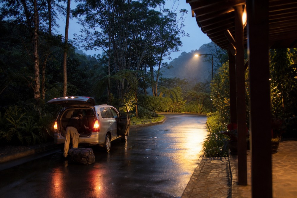 A pre-dawn hotel driveway with a vehicle's headlights on and a duffel bag being loaded, streetlight glow on wet tarmac, empty road beyond
