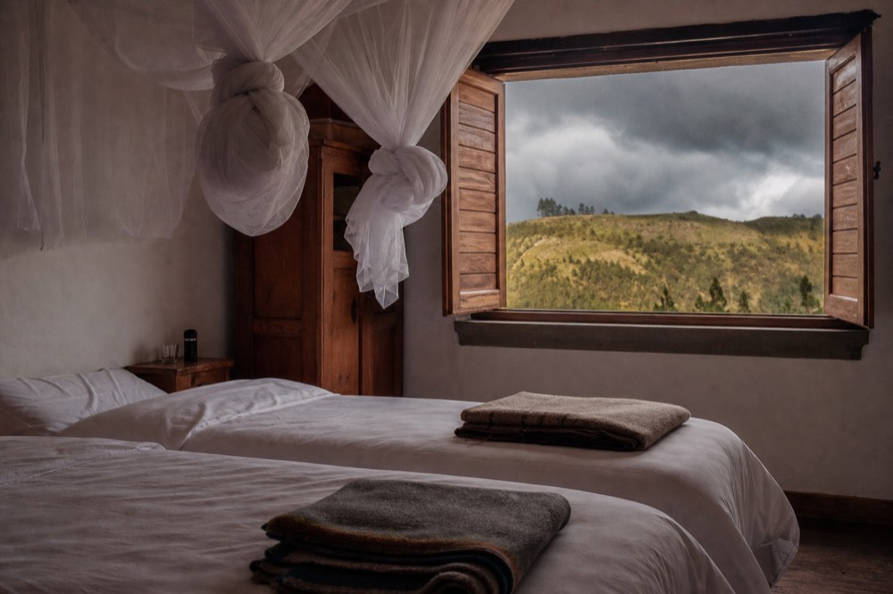 A twin room in a small guesthouse with white mosquito nets tied above the beds, folded blankets, a wooden shutter open onto green hillside