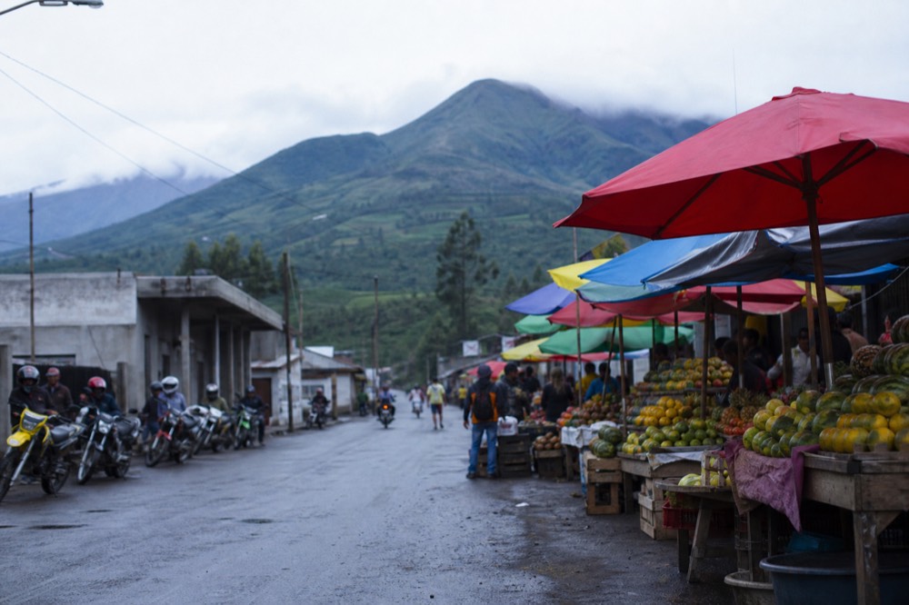 A busy small-town street in the afternoon with fruit stalls under bright umbrellas, motorbike taxis parked in a row, green volcanic slopes closing the view