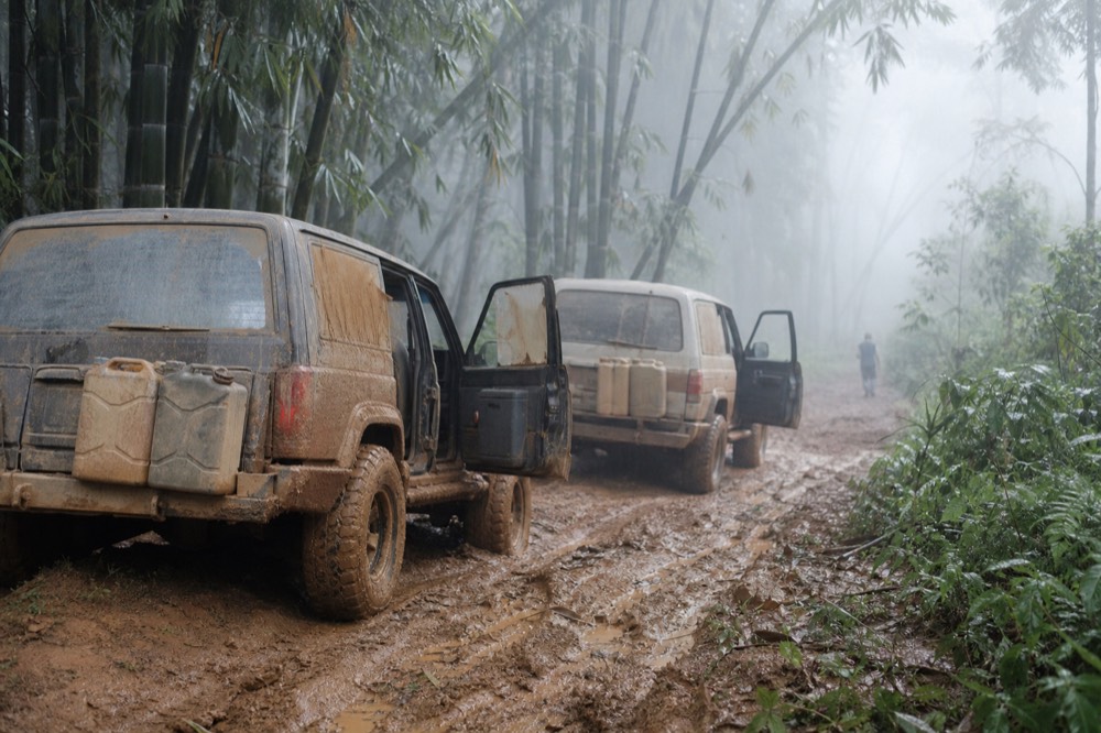 Two mud-caked 4x4 vehicles parked nose to tail on a rutted track at the forest edge, drivers' doors open, jerry cans strapped to the rear bumper