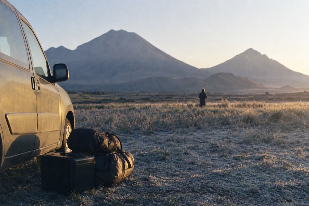 Duffel bags and a scuffed camera case stacked beside a vehicle at first light, frost-white grass underfoot, breath visible in the cold air