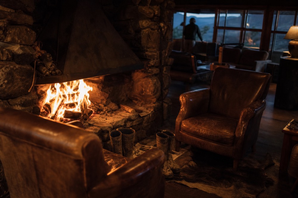 A lodge lounge at night with a log fire burning in a stone hearth, two worn leather armchairs, muddy boots drying on newspaper by the grate