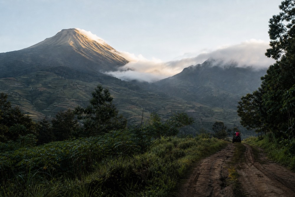 Mist pouring over a saddle between two volcanic cones at sunrise, gold light on the upper slopes, dark forest and cultivated valleys below