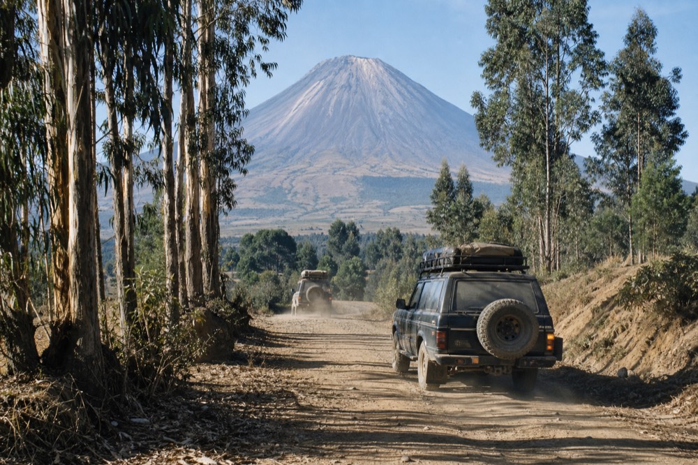 Dusty dry-season track winding between eucalyptus trees toward a bare volcanic cone, hard midday sunlight, pale cracked earth at the roadside