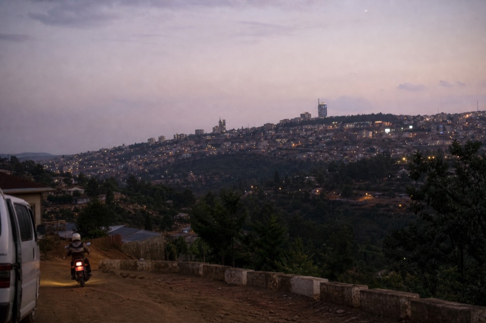 Kigali skyline at dusk seen from a hillside, low-rise buildings and streetlights spreading over green ridges, purple sky above distant hills