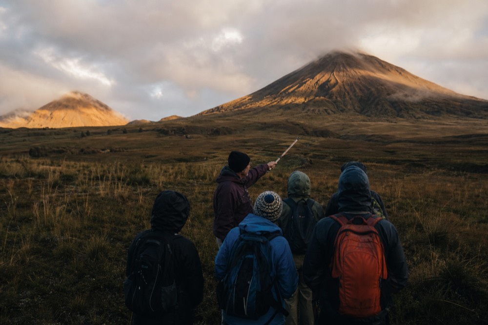 A ranger guide pointing uphill with a carved walking stick while trekkers in rain jackets listen, wet tussock grass and low cloud around them