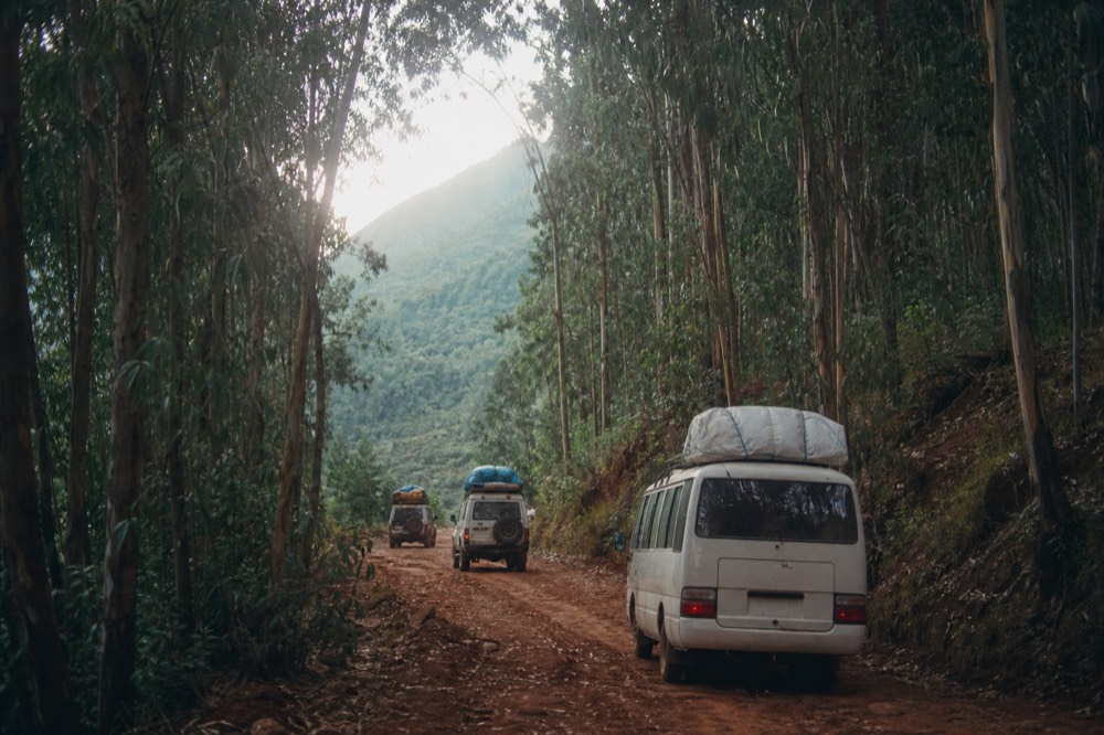 A minibus and two 4x4s on a red dirt road between tall eucalyptus trees, morning haze, green hills stepping away into the distance