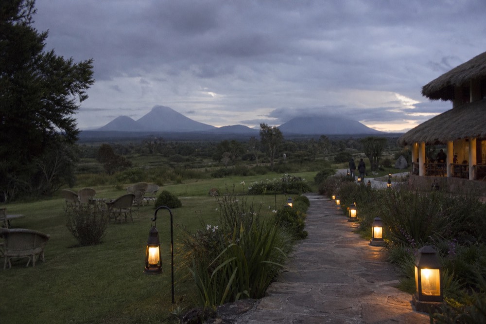 A quiet lodge courtyard at dusk with lanterns lit along a stone path, cane chairs on a lawn, volcano silhouettes low on the horizon