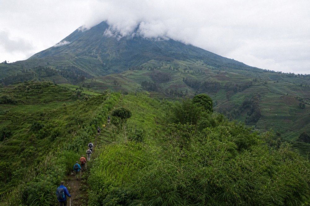 Trekkers strung out in single file climbing a green ridge above cultivated fields, seen from far behind, low cloud sitting on the volcano summit above them