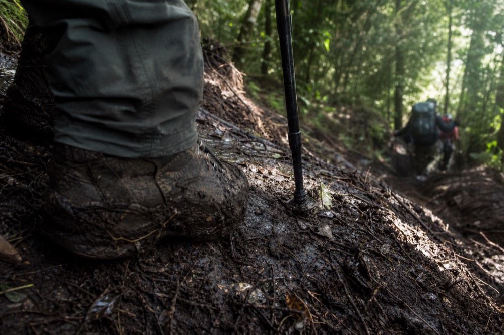 A pair of muddy boots planted on a slick descending slope of dark volcanic soil, churned earth and crushed vegetation, walking pole tip driven in beside them