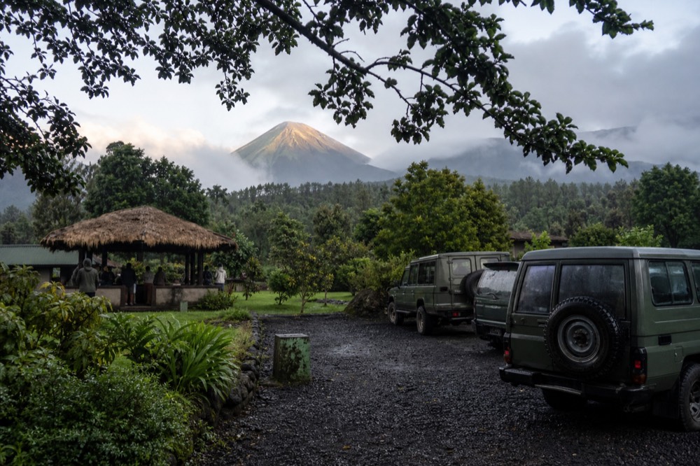 Wide view of a park headquarters courtyard at dawn with parked 4x4s, a thatched briefing shelter and volcanic peaks catching first light beyond