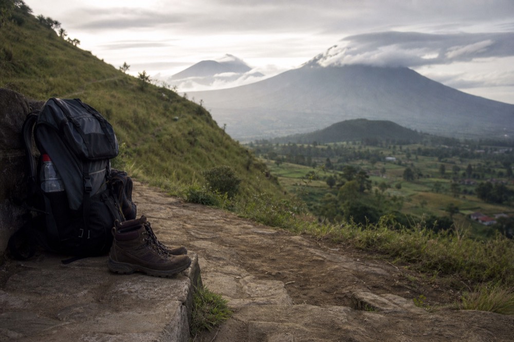 Worn hiking boots and a loaded daypack on a stone step beside a steep grassy hillside path at home, overcast light, training ground rather than jungle