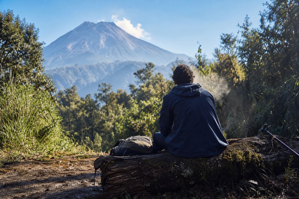 Trekker resting on a mossy log at a forest clearing edge, back to camera, steam rising from damp clothing, green wall of vegetation behind