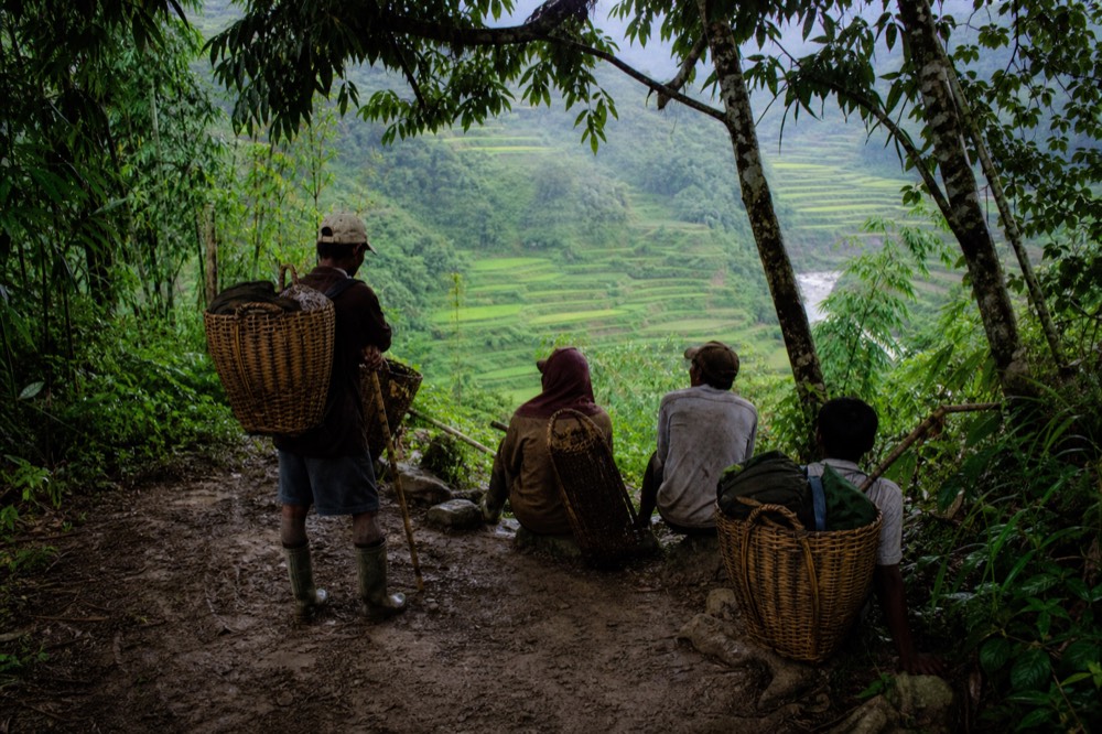 Local porters waiting at a trailhead with woven baskets and walking sticks, mud-spattered boots, terraced green fields dropping away behind them