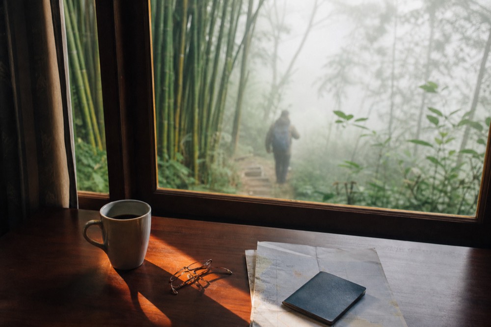 A passport, folded travel insurance paperwork and a pair of reading glasses on a wooden hotel desk beside a mug of black coffee in warm morning light