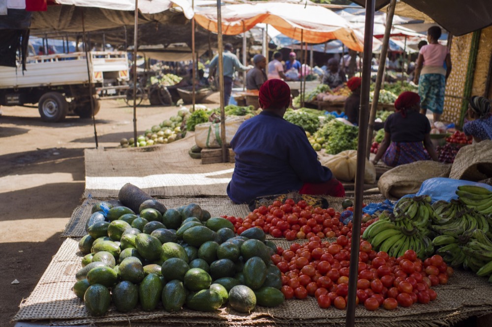 An open-air Kigali produce market at midday with pyramids of tomatoes, avocados and green bananas on woven mats under bright canvas awnings
