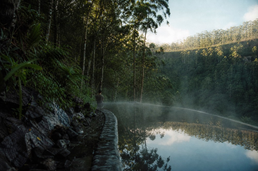 An infinity-edge pool cut into black volcanic stone, steam rising from the water at dawn, a green crater rim and eucalyptus stand mirrored on the surface