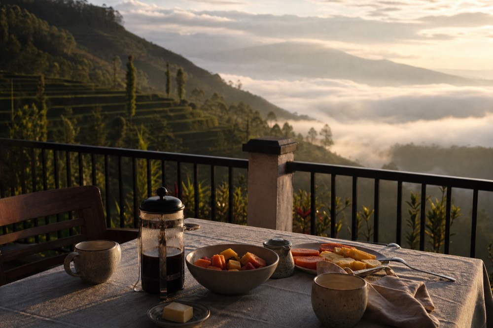 Breakfast laid on a terrace table at sunrise, coffee pot steaming beside fresh fruit and a linen napkin, mist filling the valley below the railing