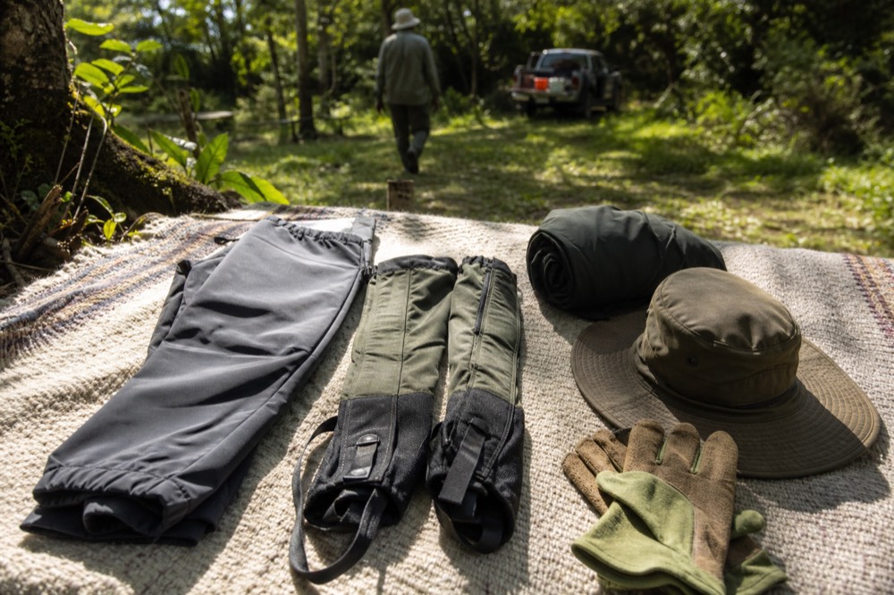Clean waterproof trousers, gaiters and gardening gloves laid out on a woven bedspread beside a wide-brimmed hat and a rolled rain shell