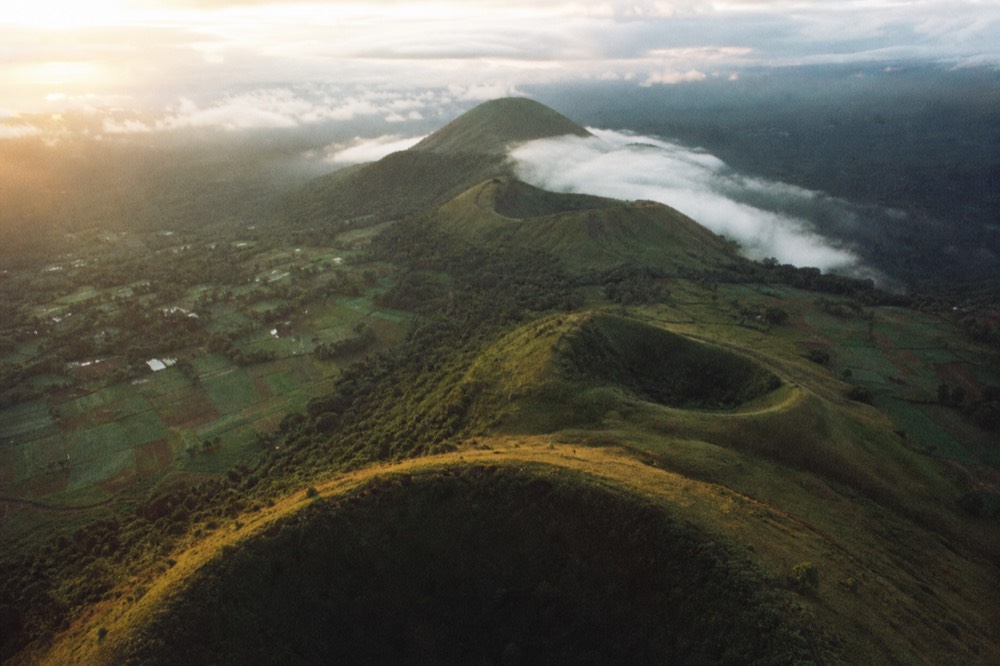 Aerial view along a chain of volcanic cones at golden hour, cloud pooling in the saddles between them, patchwork farmland running to the forest boundary