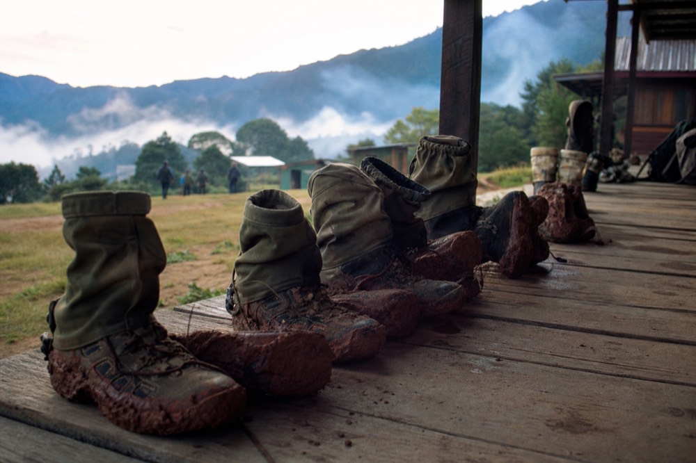 A row of muddy hiking boots and canvas gaiters drying on a wooden lodge veranda in morning light, red earth caked thick on the soles