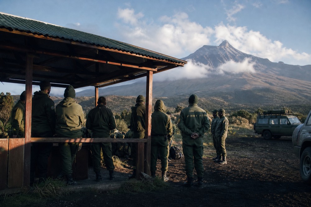 Park rangers in green uniforms standing in a loose semicircle at an open-sided briefing shelter at first light, volcano slopes wrapped in cloud behind them