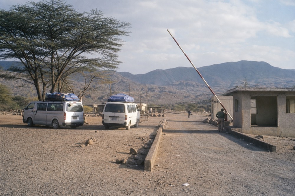 A quiet East African land border post at midday, striped barrier arm raised, small concrete immigration building and parked minibuses under a dusty acacia