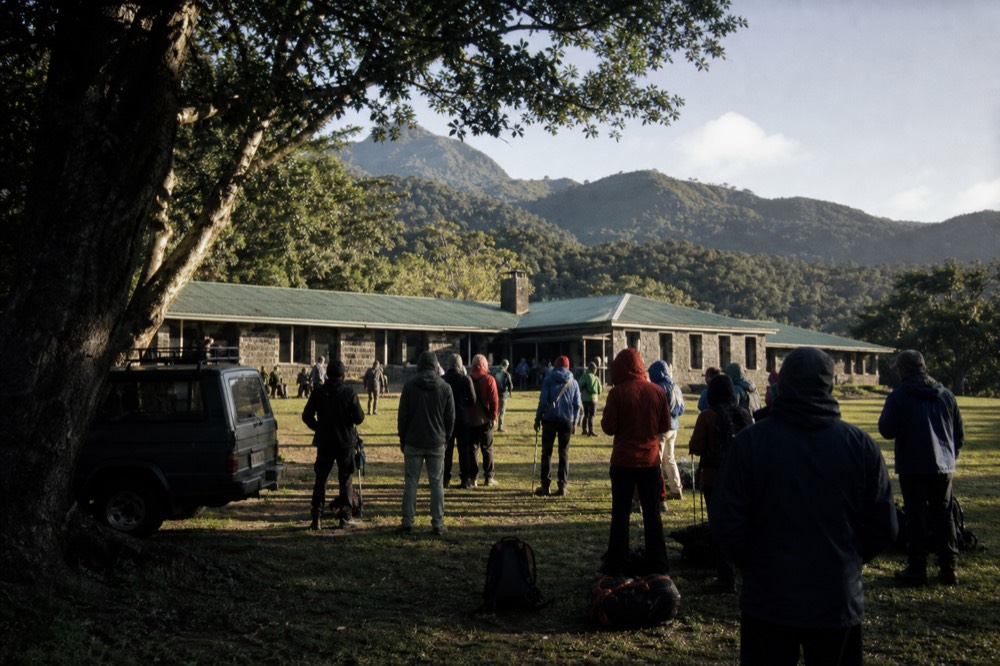 Trekkers in rain jackets gathered on a lawn outside a low stone park headquarters building at sunrise, green hills rising behind them, no signage visible