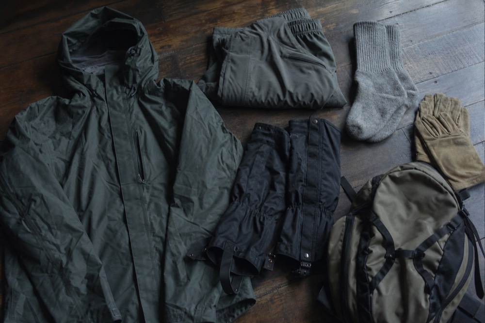 Flat-lay of trekking kit on a wooden floor: waterproof jacket, folded trousers, wool socks, canvas gloves, gaiters and a small daypack, soft window light
