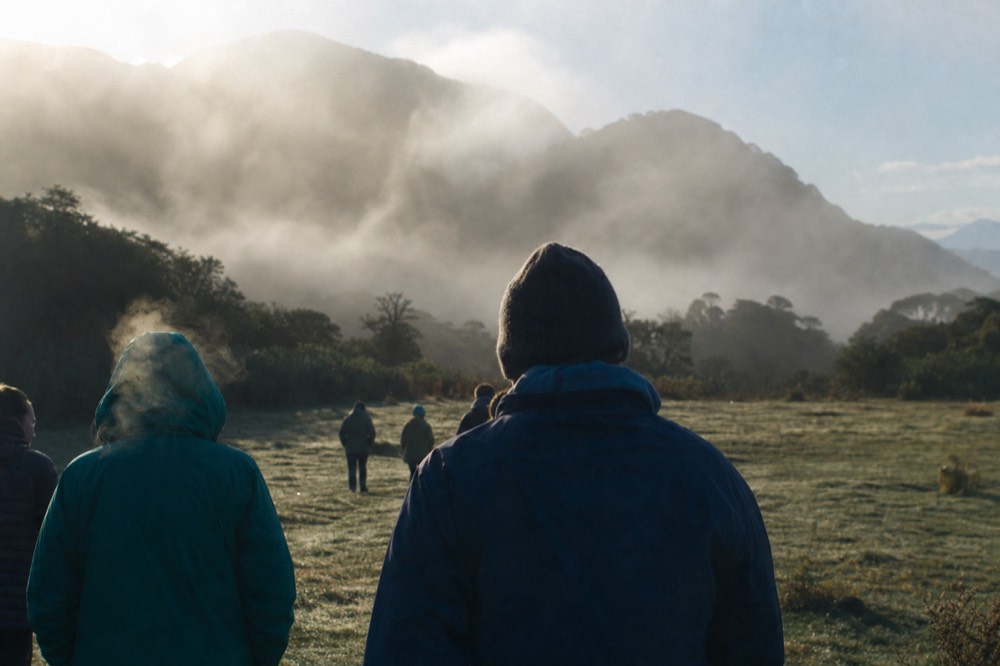 Trekkers in fleeces gathered on a misty grass field at first light with hills behind, breath visible in cold air, seen from behind so no faces show