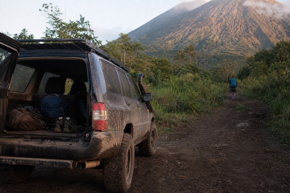 Mud-splattered 4x4 vehicle parked at a forest trailhead with the rear door open showing boots and packs, green volcanic slope rising behind